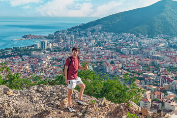 Young adult man showing thumbs up while standing on top of a mountain with the city rooftops of Budva and the Adriatic Sea skyline in the background, Montenegro. Hiking on the Budva Riviera in summer