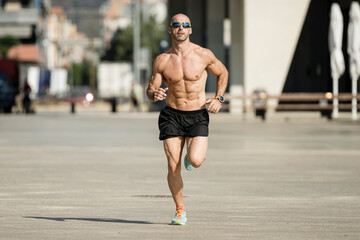 Athletic young man running in the street wearing sunglasses. Front view of sportsman jogging outdoors shirtless.