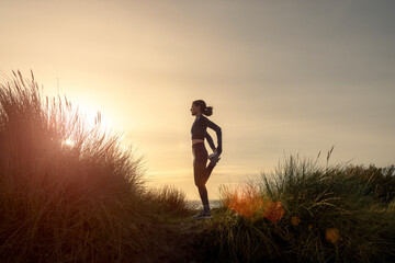 Sporty woman stretching her legs, doing standing quadricep front thigh stretch outdoors at sunrise - obrazy, fototapety, plakaty