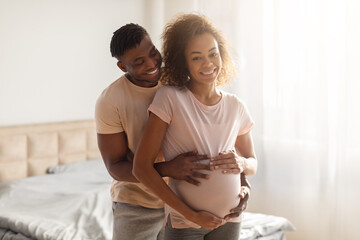 African husband tenderly hugging his pregnant wife's belly in bedroom