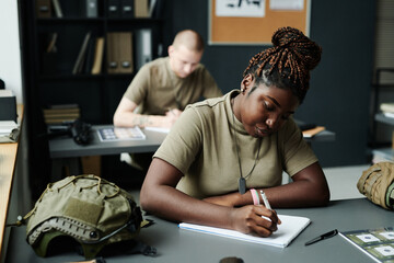 Young African American female student making notes in notepad after teacher while sitting by desk...