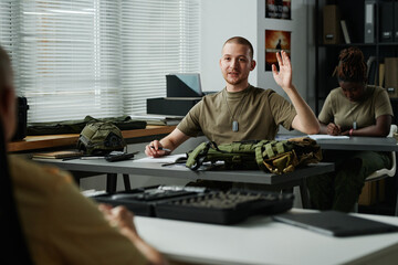 Young male student raising hand and looking at teacher at lesson while asking him question after lecture about military training - obrazy, fototapety, plakaty
