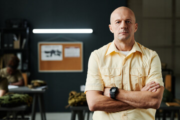 Bald serious male teacher of military training crossing arms by chest and looking at camera while standing against classroom interior - Powered by Adobe