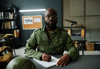 Young serious African American man in camouflage uniform sitting by desk, making notes and...