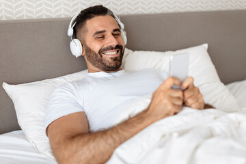 man lying in bed with earphones and phone in bedroom - obrazy, fototapety, plakaty