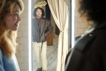 Reflection in mirror of young man trying on stylish leather jacket in fitting room in boutique and consulting with his wife or girlfriend - obrazy, fototapety, plakaty