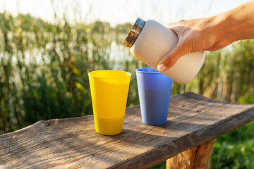 A hand, pouring tea from a white thermos into a blue cup on a wooden table with a yellow cup next...