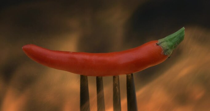 Super slow motion macro of fresh bio organic red hot spicy chili pepper on fork is burning in fire orange flames with smoke at 1000 fps isolated on dark background.