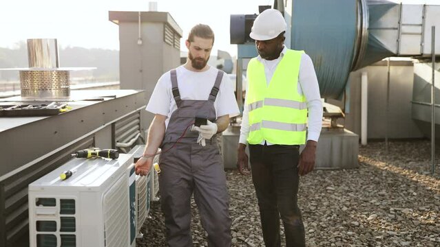 Focused technician standing and using multimeter to air conditioner while male supervisor controlling process. Multicultural repairmen measuring voltage and monitoring operation of device on factory.