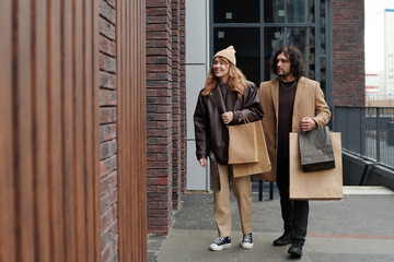 Young shoppers in stylish casualwear carrying paperbags with purchases while walking along shop...