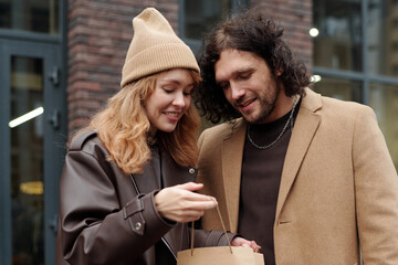 Pretty woman in stylish casualwear opening paperbag with purchase and boasting to her boyfriend in urban environment after shopping