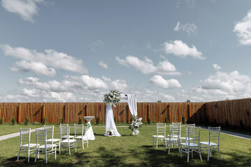 Outdoor wedding venue featuring white chairs and a floral arch on a green lawn. A wooden fence and...