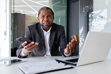 Portrait of a worried African American businessman sitting in the office at a desk, holding a...