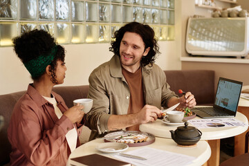 Biracial male and female colleagues having breakfast at cafe discussing charts on laptop screen