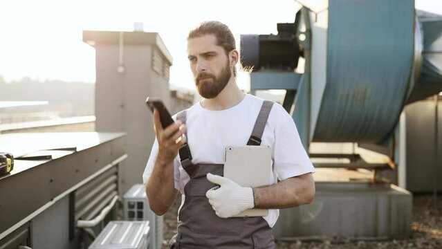Serious factory worker with braided hair holding tablet while having conversation on wireless mobile phone. Caucasian qualified man standing and resolving issues related to devices on factory roof.