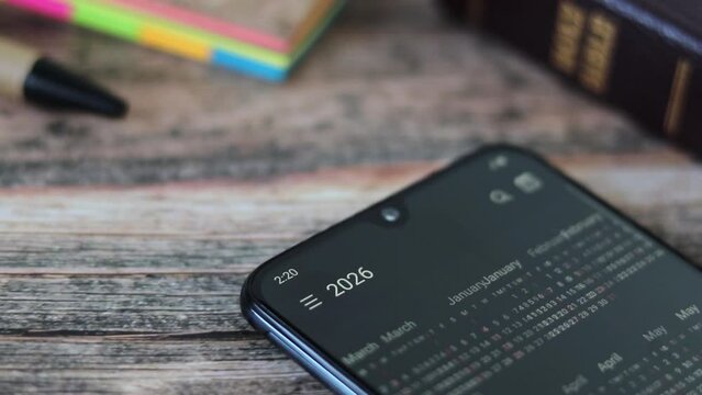 Digital calendar on smartphone screen with Christian holy book in the background. Close-up. Time running, daily bible study, change, progress, biblical concept.