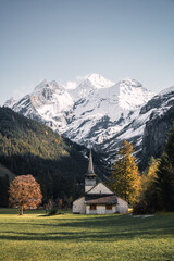 Fototapeta na wymiar Autumn landscape view of a typical alpine church with colourful trees and snow-capped mountains in the background