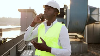 Portrait of male worker dressed in work uniform standing and smiling while talking on digital mobile on fresh air. African american man having conversation and working with devices on roof of factory.