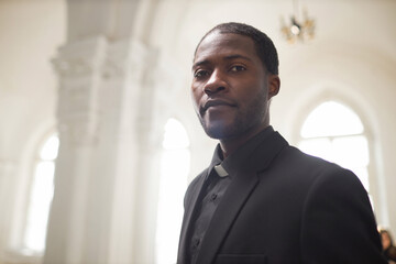 Portrait of young Black man as religious priest looking at camera in ethereal church setting, copy...