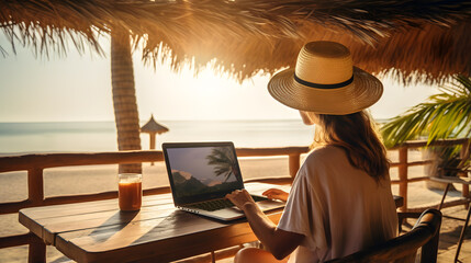 Woman wearing a straw hat working remotely with her laptop from a tropical beach, facing the ocean, under the shade of a thatched roof in the golden hour. - obrazy, fototapety, plakaty