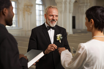 Waist up portrait of smiling senior groom exchanging rings with wife during wedding ceremony at...
