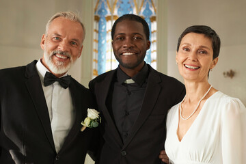 Waist up portrait of newlywed senior couple posing with smiling priest during wedding ceremony in...