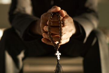 Closeup of priest holding wooden rosary beads and praying in ethereal sunlight, copy space - obrazy, fototapety, plakaty