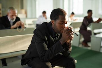 Portrait of Black young man in silent prayer at church holding rosary beads in sunlight