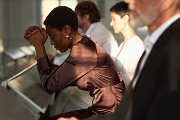 Side view portrait of Black adult woman praying in church during Sunday meeting lit by sunlight