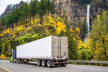 Blue and green classic big rig semi truck transporting cargo in reefer semi trailer moving on the highway road driving past a mountain range with autumn forest and Multnomah Falls