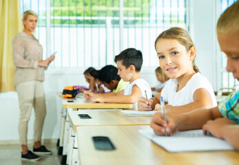 Portrait of positive tween schoolgirl sitting on lesson in classroom, looking at camera with smile