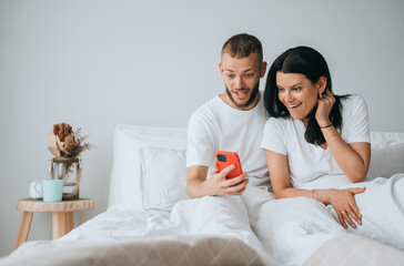 Obraz na płótnie Canvas Amazed couple sitting in bed looking at phone with surprised face expression, shocked by great news. Mockup with empty space on background. Sale, discount. Furniture ad.