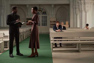 Side view portrait of African American pastor advising woman after Sunday service in catholic...