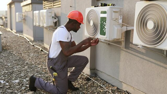 Qualified builder wearing uniform inspecting air conditioner and using screwdriver during repairing work. Black serious male standing on one knee and fixing device in roof of building.