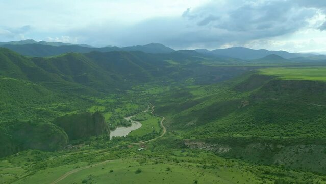 An aerial view of high rocky mountains covered with green grass and trees on an overcast summer day. Small villages near the river with country roads. Dzoraget River, Canyon, Armenia. Drone footage