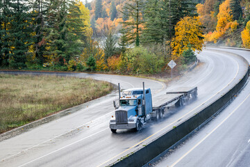 Light blue classic big rig bonnet semi truck tractor transporting two empty flat bed semi trailer driving on the wet autumn highway road with rain dust at rainy weather - obrazy, fototapety, plakaty