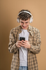 Fototapeta premium One man caucasian young male stand studio shot on beige background use mobile phone smartphone with headphones listen music send messages texting or browse internet online app copy space