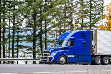 Fototapeta na wymiar Streamlined shape blue big rig semi truck with grille guard transporting cargo in refrigerator semi trailer driving on the road along the river with autumn trees line