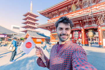 Handsome young tourist enjoying summer holiday in Tokyo, Japan - Traveling life style concept with smiling man taking selfie on city street with japan flag- Tourism and summertime vacation concept - obrazy, fototapety, plakaty