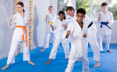Concentrated black teenager in kimono practicing punches in gym during group martial arts workout....