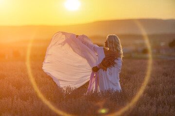 Blonde woman poses in lavender field at sunset. Happy woman in white dress holds lavender bouquet. Aromatherapy concept, lavender oil, photo session in lavender - obrazy, fototapety, plakaty