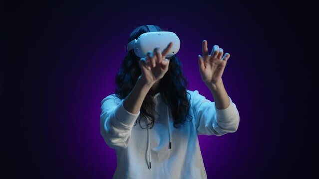 A happy girl in virtual reality glasses interacts with the virtual world using her fingers. She is in a room with a dark purple background illuminated by blue light. High quality 4k footage