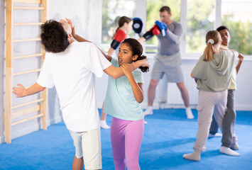 Fototapeta premium Girls and boys practicing in pair self-defence movements with male trainer supervision