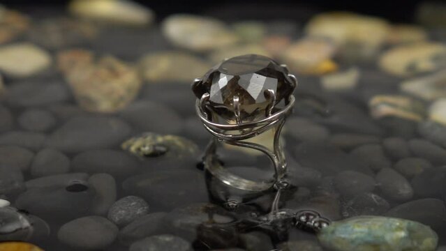 Drops of water fall on the silver ring with smoky topaz on a background of water and black pebbles, slow motion