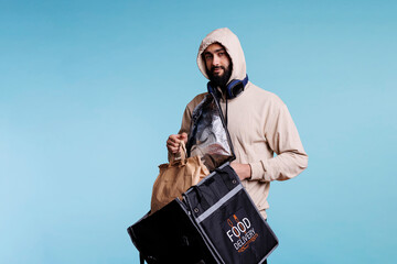 Young arab man taking paper bag with food from backpack and looking at camera. Deliveryman in hood...