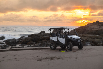 Rescue ATV buggy car on sandy beach at sunset hour. Bright orange sky and big ocean waves in the...