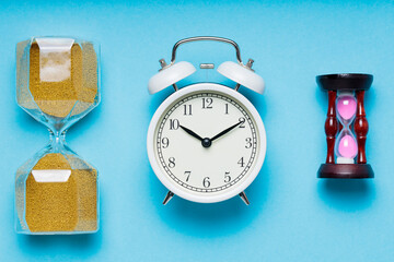 Two Hourglasses and White Alarm Clock on Blue Background, Time Concept