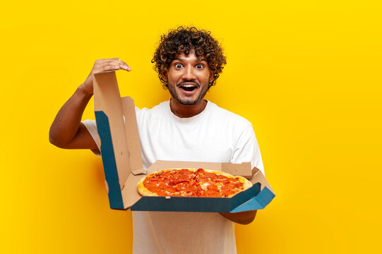 hungry Hindu guy opens a box with delicious pizza on a yellow isolated background, young Hindu man holds fast food