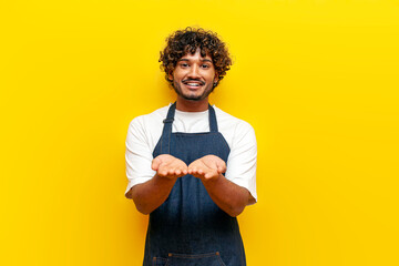 young surprised guy Indian waiter in an apron begs on a yellow isolated background