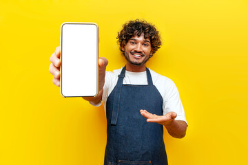 young guy Indian waiter in apron shows blank screen of smartphone on yellow isolated background